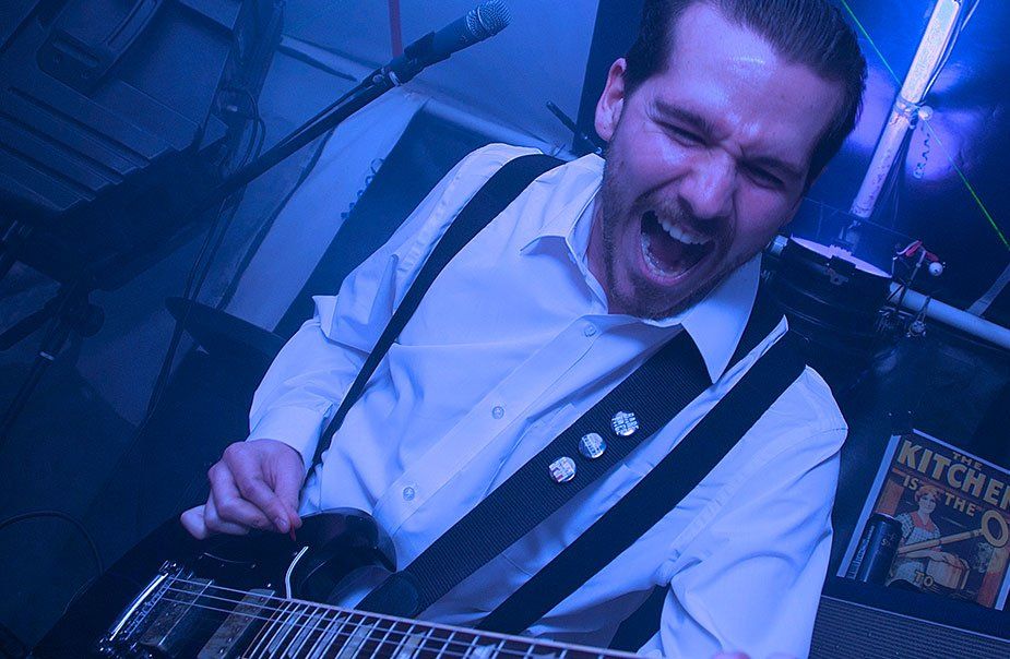 A man in a white shirt and suspenders is playing a guitar in front of a microphone.