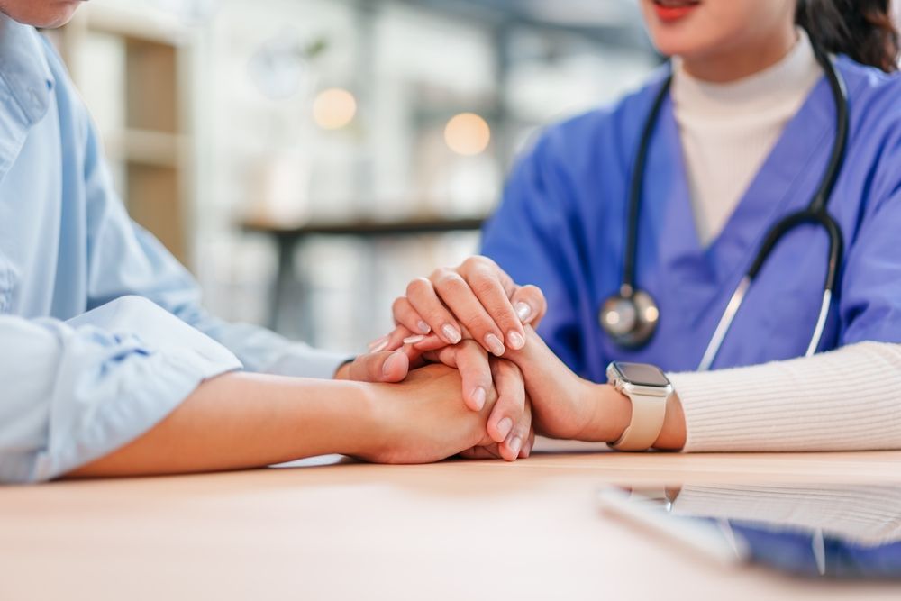 A healthcare worker in blue scrubs holds a patient's hand, offering support.