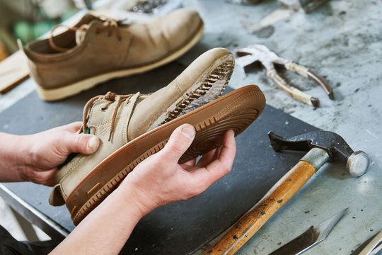 Hands repairing a worn beige shoe on a workbench with a hammer nearby Hands repairing a worn beige shoe on a workbench with a hammer nearby