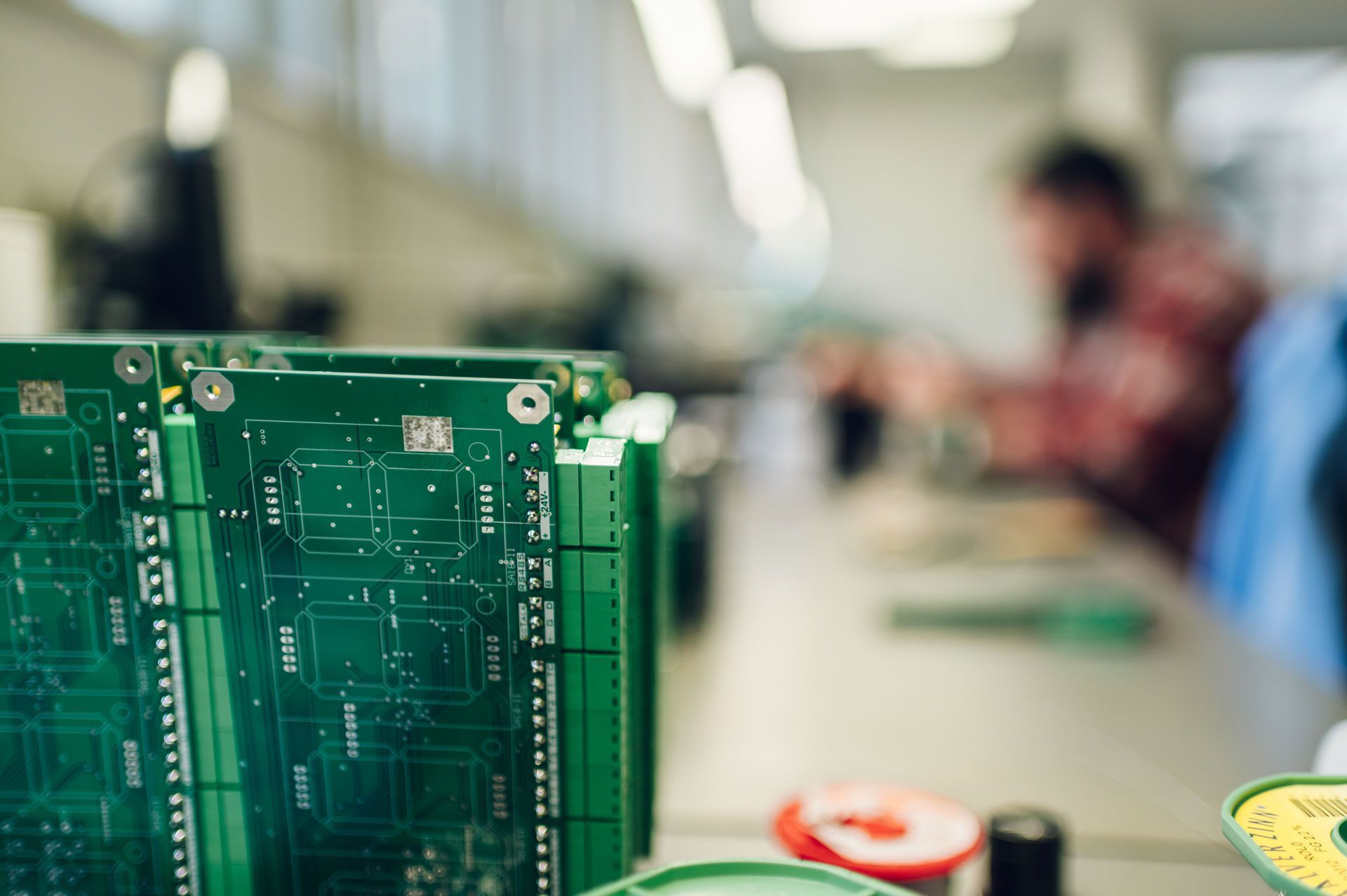 Green circuit boards stacked in focus, with a blurred person soldering in the background.
