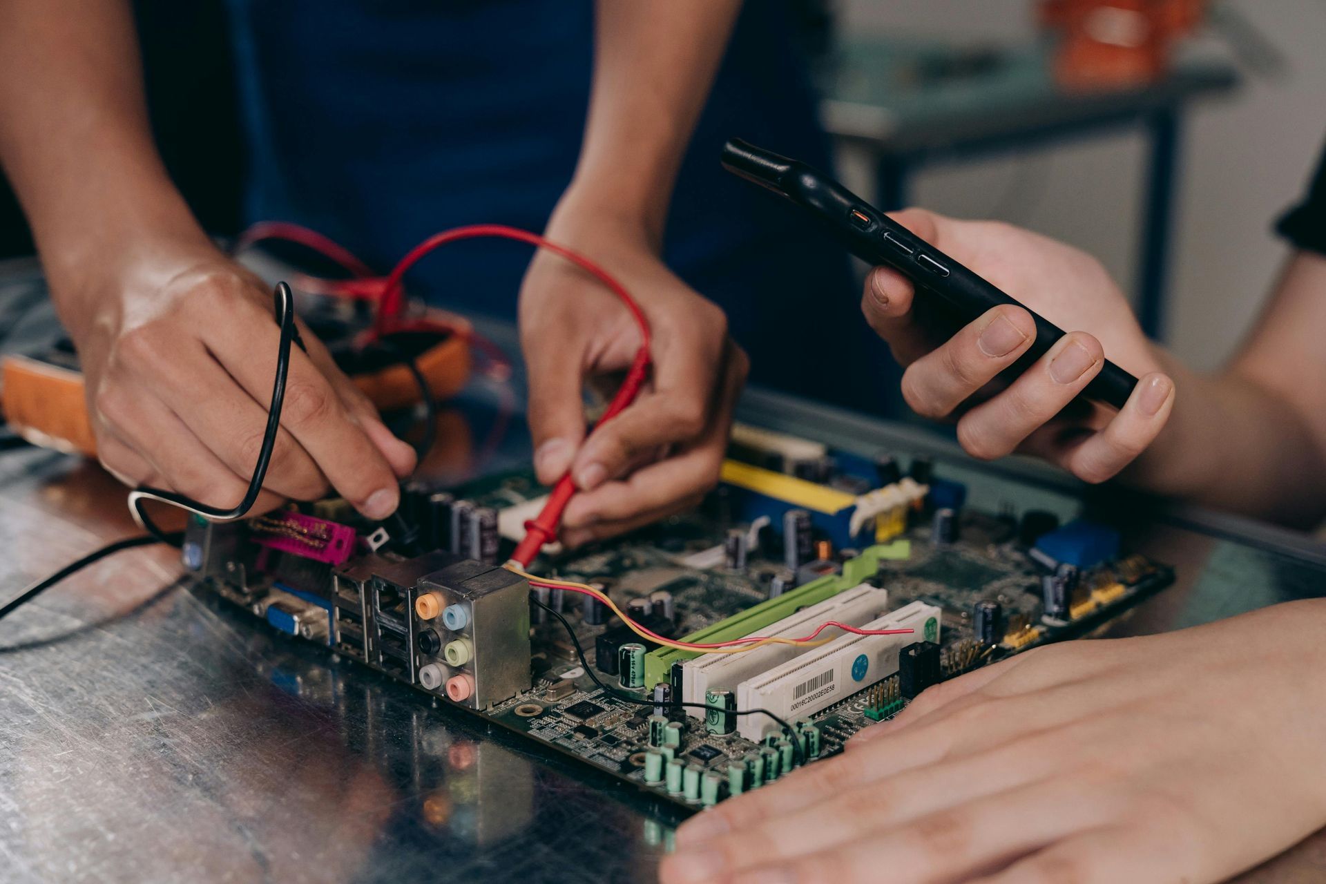 Hands testing a circuit board with probes and a multimeter on a metal table.