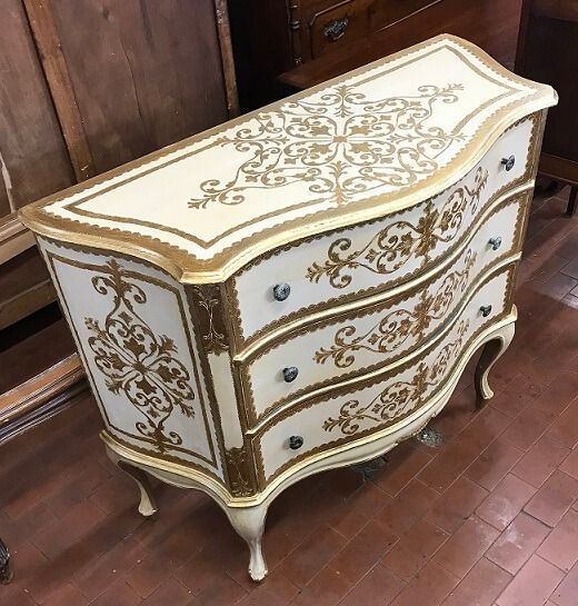 Comò antico color crema con finiture dorate e decorati motivi floreali. Tre cassetti con gambe curve, su pavimento in legno.