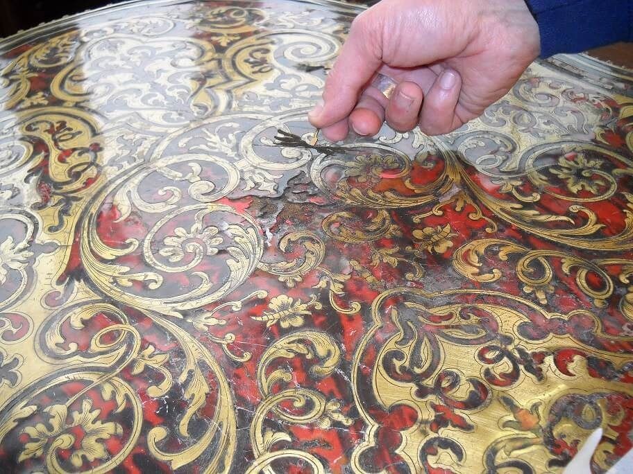 Mano che regge un frammento sopra un tavolo rosso e oro decorato e danneggiato.