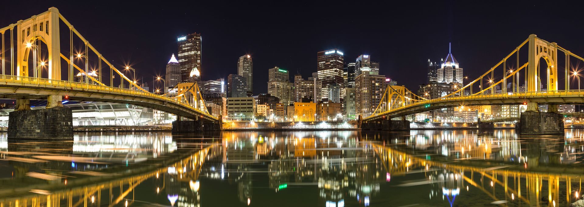 Nighttime panoramic view of a city skyline with bridges over a body of water; lights are reflected.