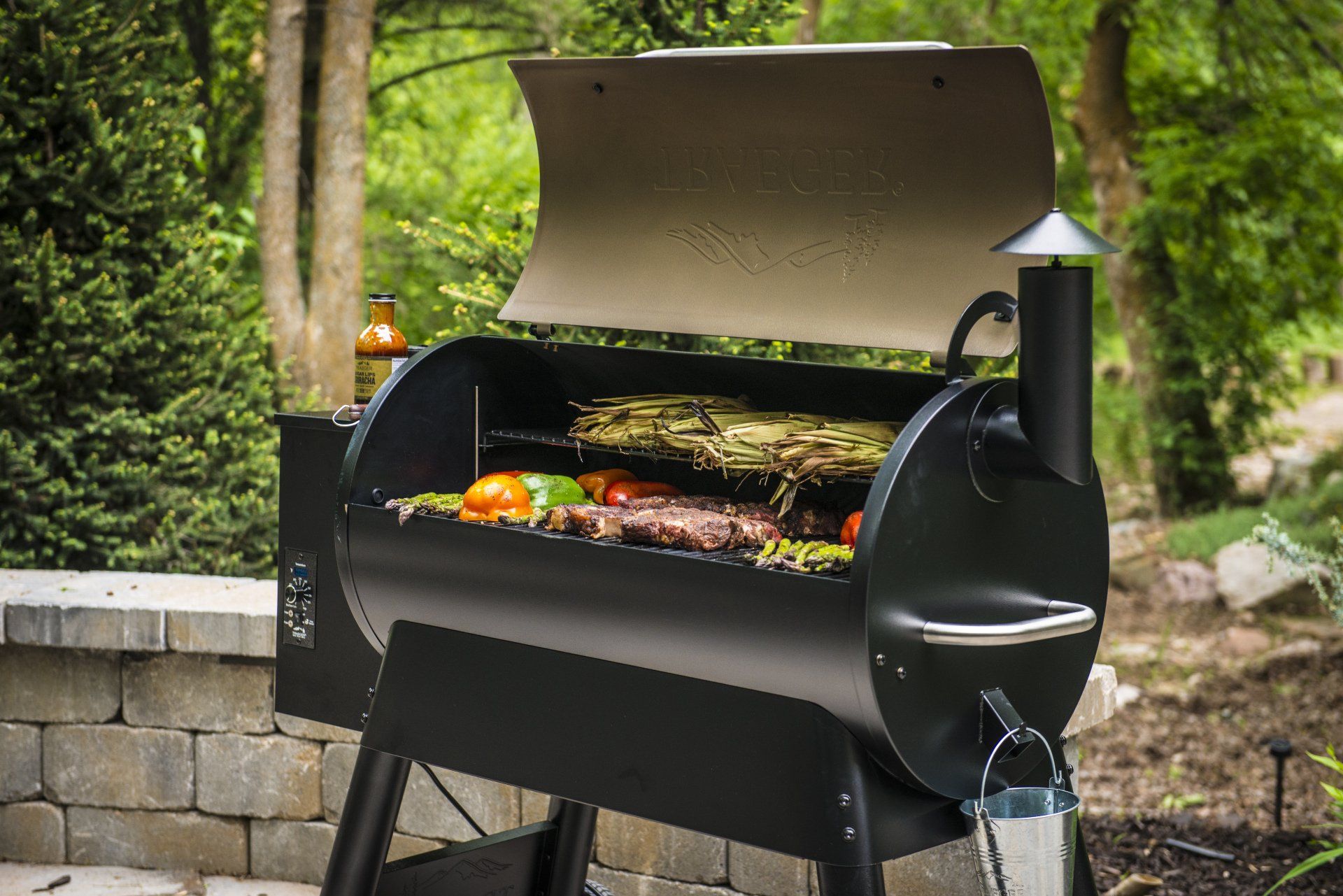 Un barbecue à pellets noir, couvercle ouvert, en train de cuire des légumes et de la viande, installé à l'extérieur sur une terrasse en pierre.