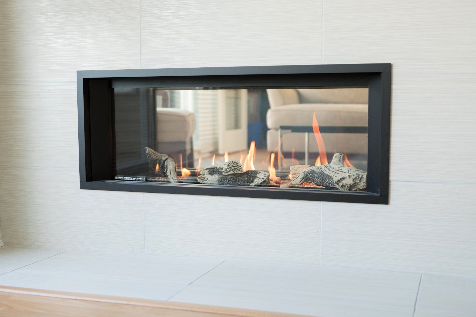 Une cheminée à gaz linéaire horizontale moderne, avec flammes, pierres et une façade vitrée, intégrée dans un mur carrelé blanc.