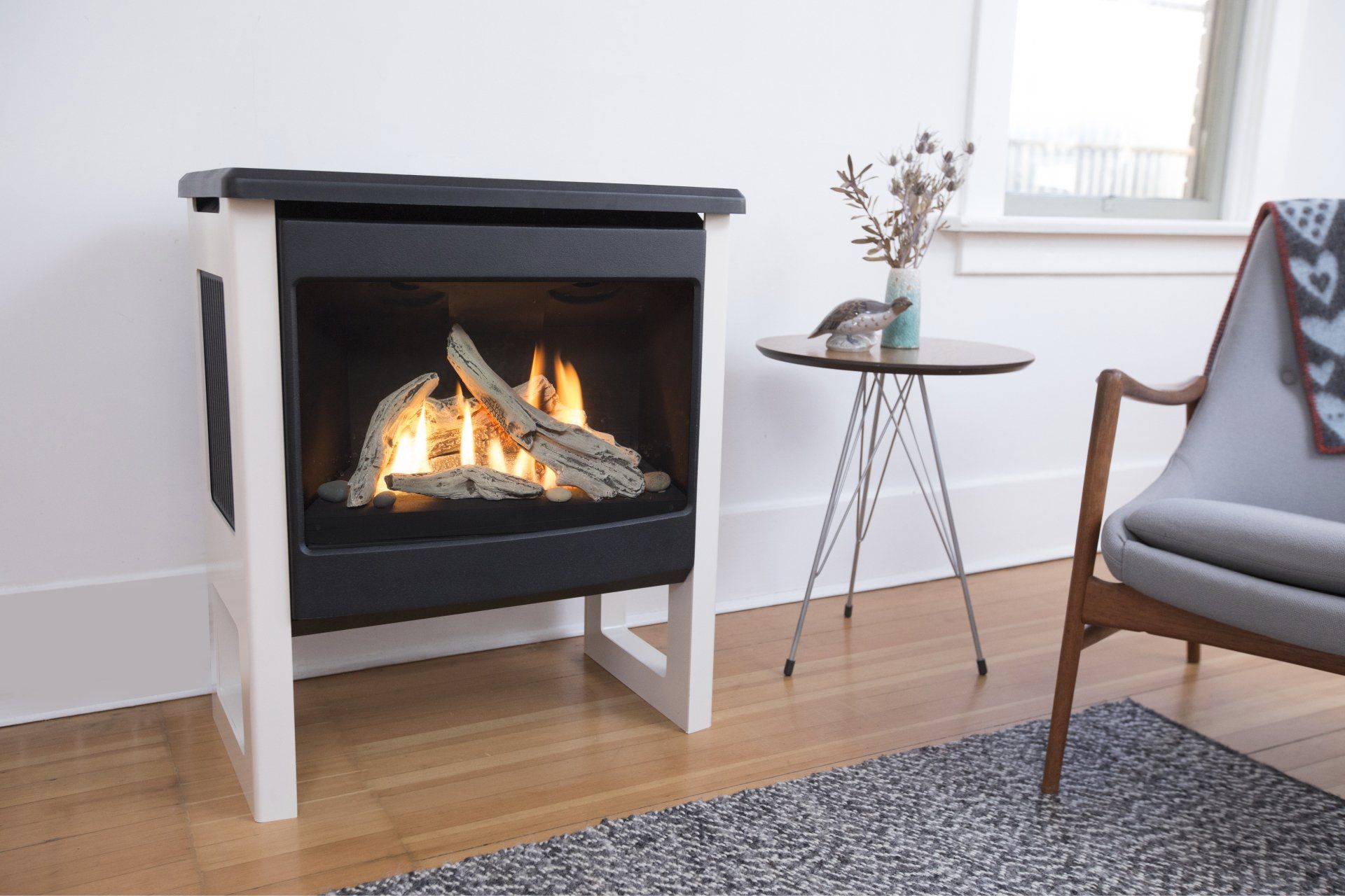 Une cheminée à la façade noire et au cadre blanc brûle. Une petite table et un fauteuil se trouvent à proximité, dans une pièce bien éclairée.