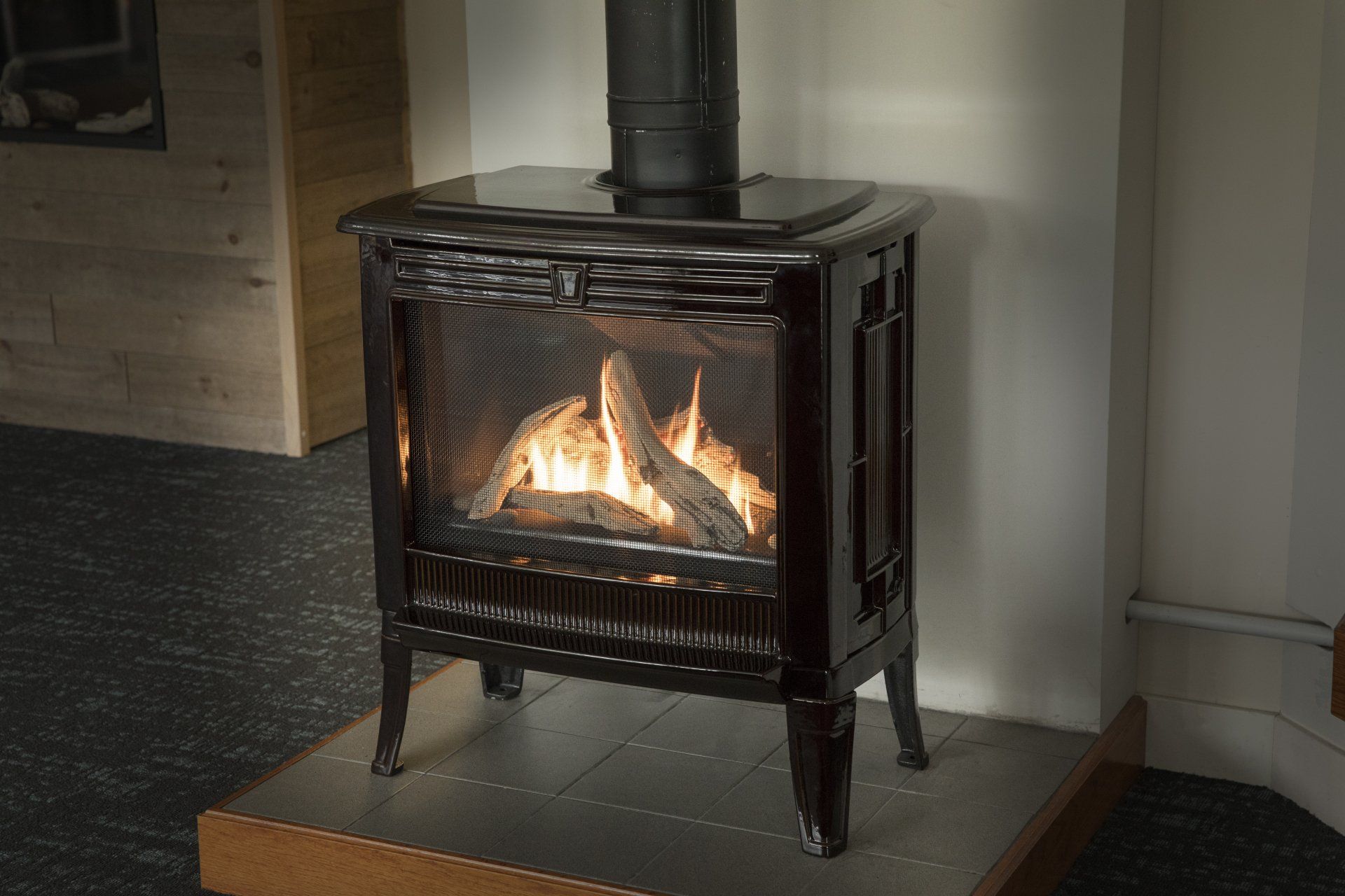 Une cheminée à gaz brun foncé avec une flamme brûlante, un tuyau de cheminée noir et une façade en verre à motifs, placée contre un mur blanc.