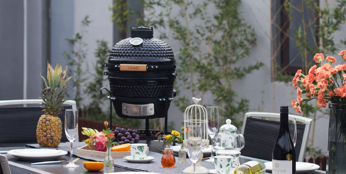 Un barbecue noir de type kamado trône sur une table dressée pour un repas, agrémentée de fruits, de verres et de fleurs, dans un cadre de terrasse.