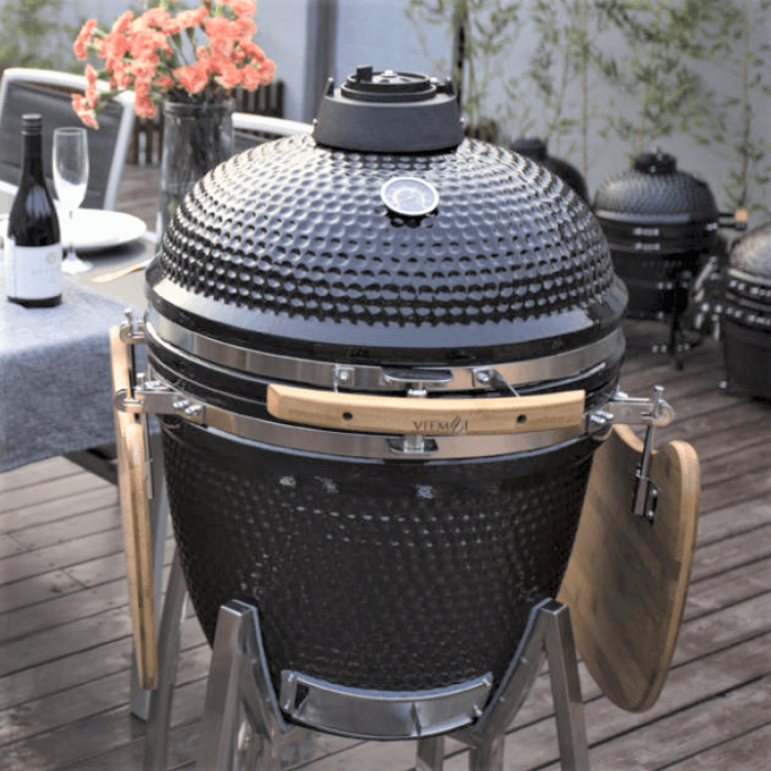 Un barbecue kamado en céramique noire, fonctionnant au charbon de bois et doté d'étagères latérales en bois, est installé sur une terrasse près d'une table à manger extérieure.