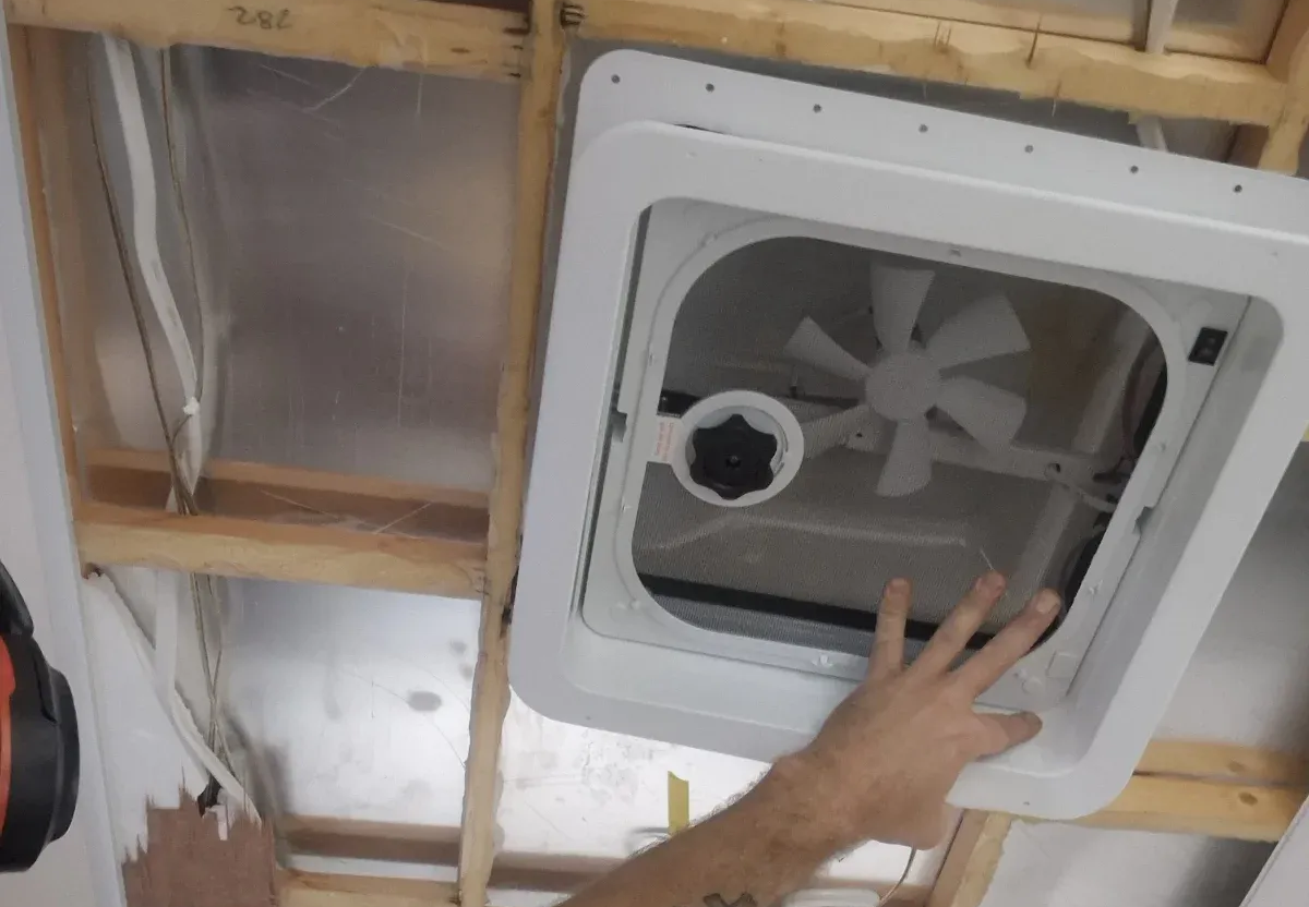 A hand holds a white vent fan inside a wood-framed opening. The fan has a central, white fan blade — Revive Caravan Repairs in Kippa-Ring, QLD