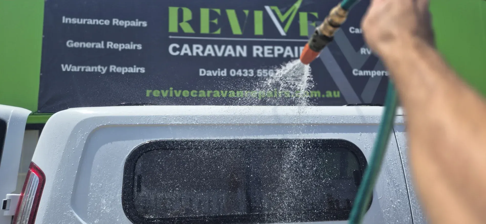 A person washing a white vehicle with a hose. Revive Caravan Repairs sign in the background — Revive Caravan Repairs in Kippa-Ring, QLD