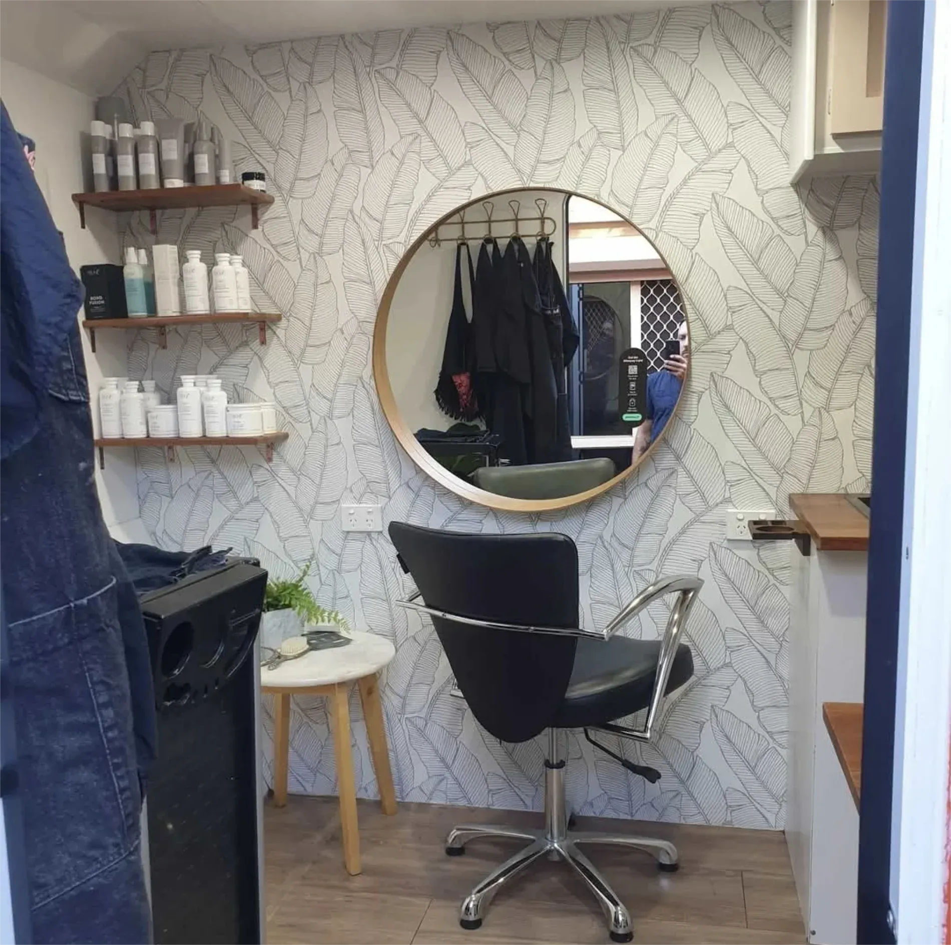 Hair salon interior with chair, round mirror, shelves with products, and leaf pattern wallpaper — Revive Caravan Repairs in Kippa-Ring, QLD