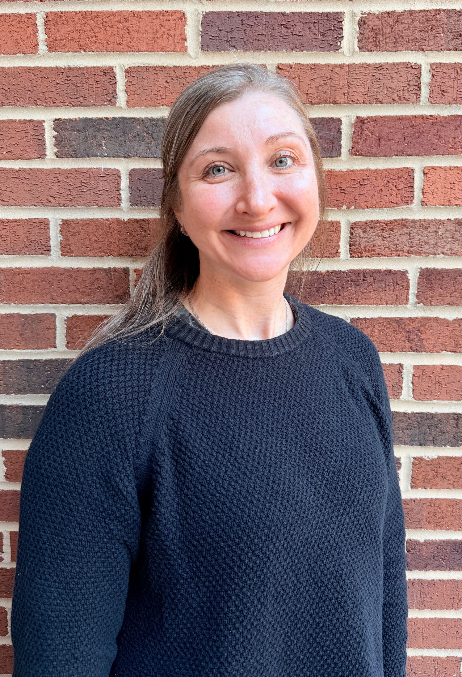 A woman with long red hair is smiling in front of a brick wall.
