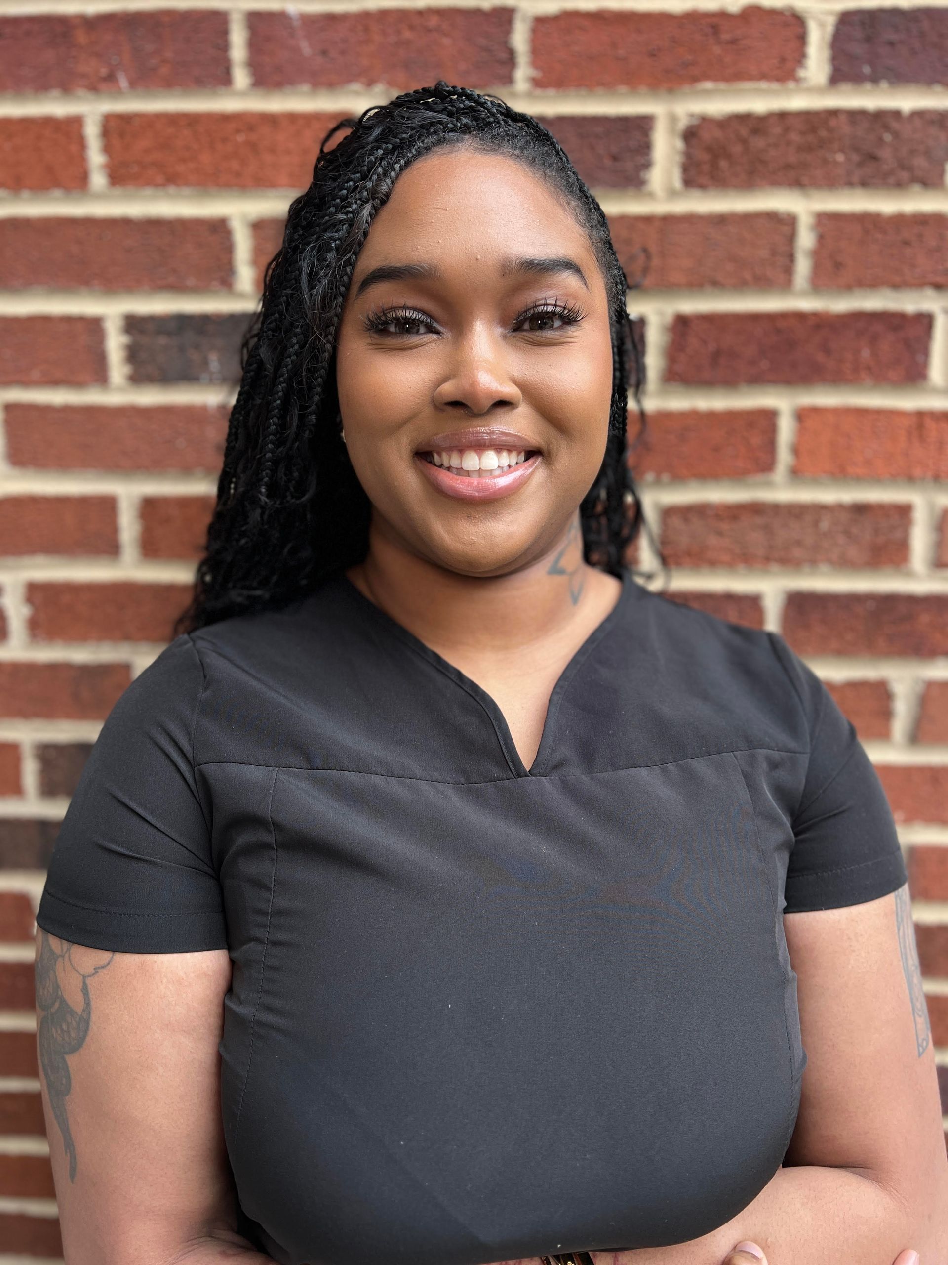 A woman in a black scrub top is smiling in front of a brick wall.