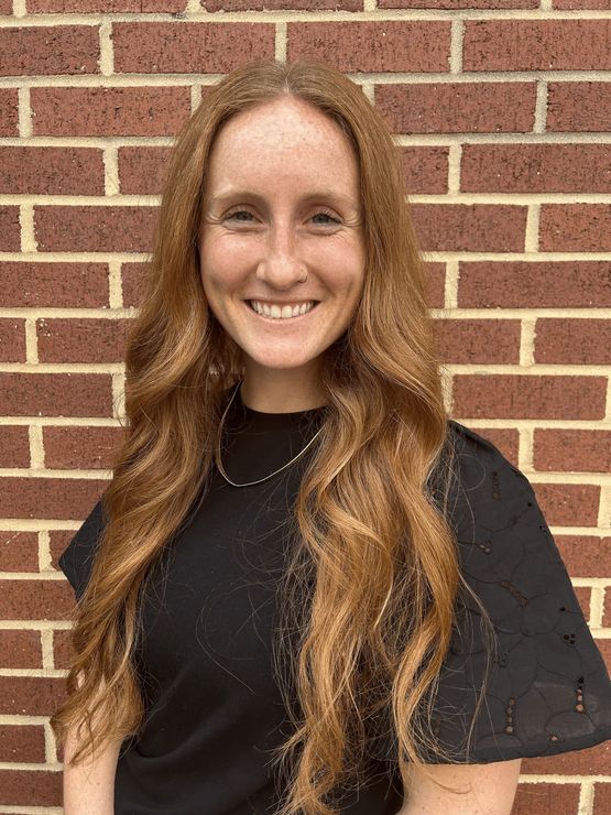 A woman with long red hair is smiling in front of a brick wall.