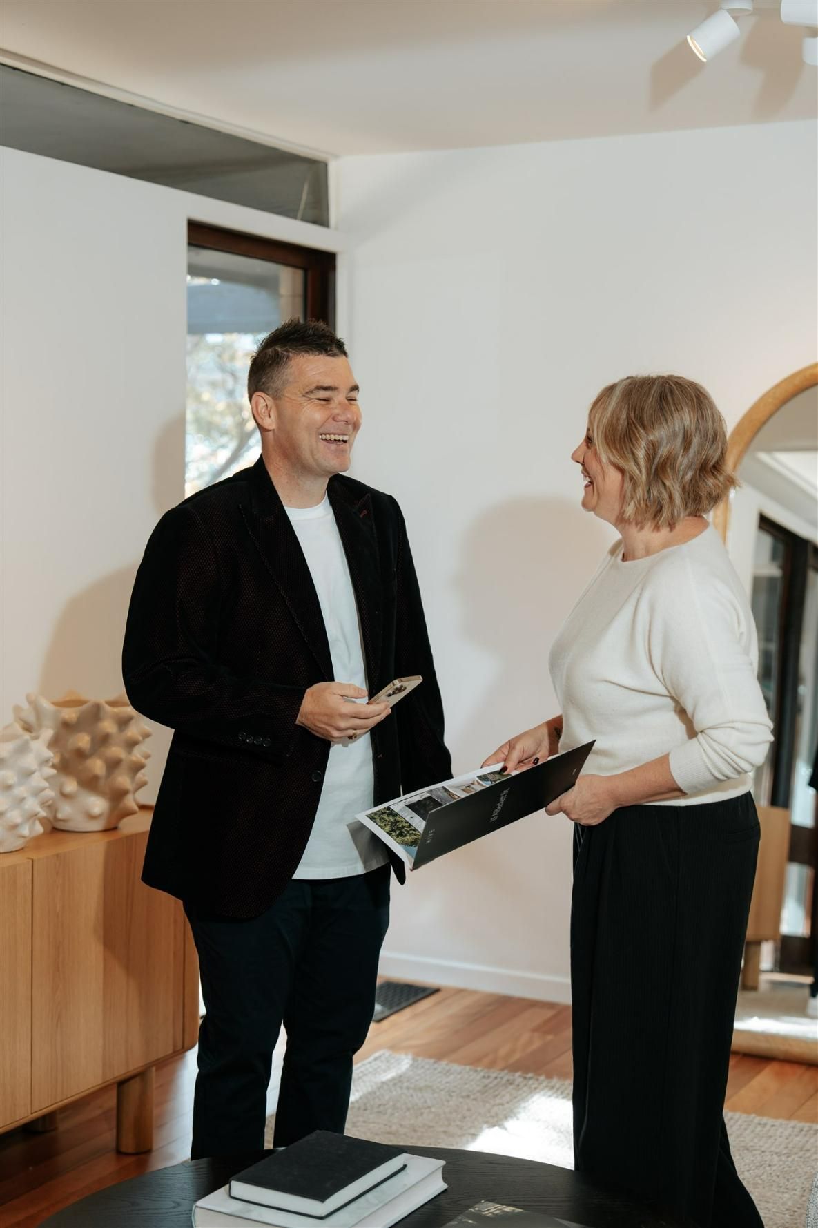 A man holding a phone is talking to a woman holding a pamphlet — Hyde Burchell Buyers Agents In Canberra, ACT