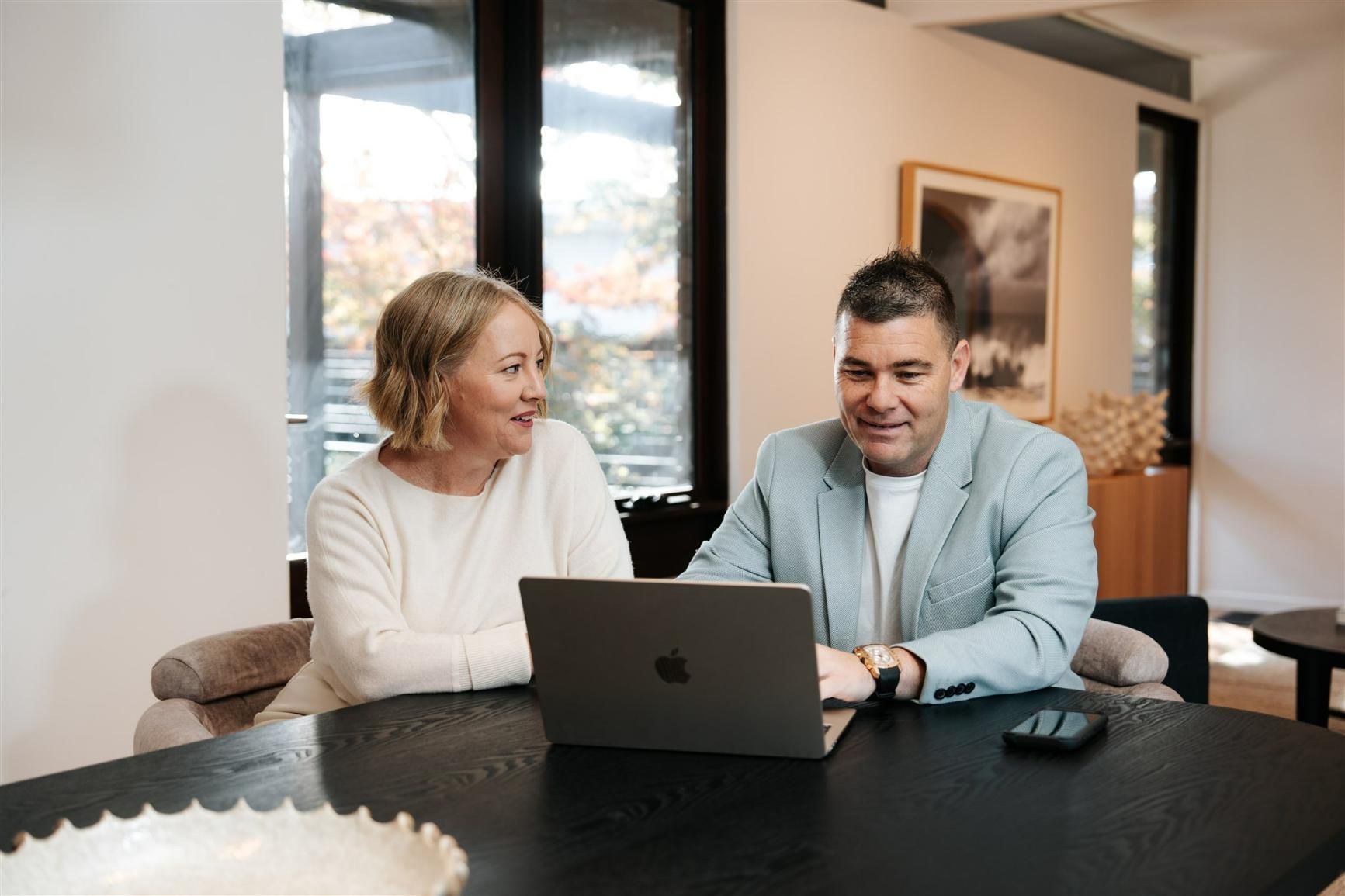 A Man and a Woman Are Sitting at a Table Looking at a Laptop Computer — Hyde Burchell Buyers Agents In Canberra, ACT