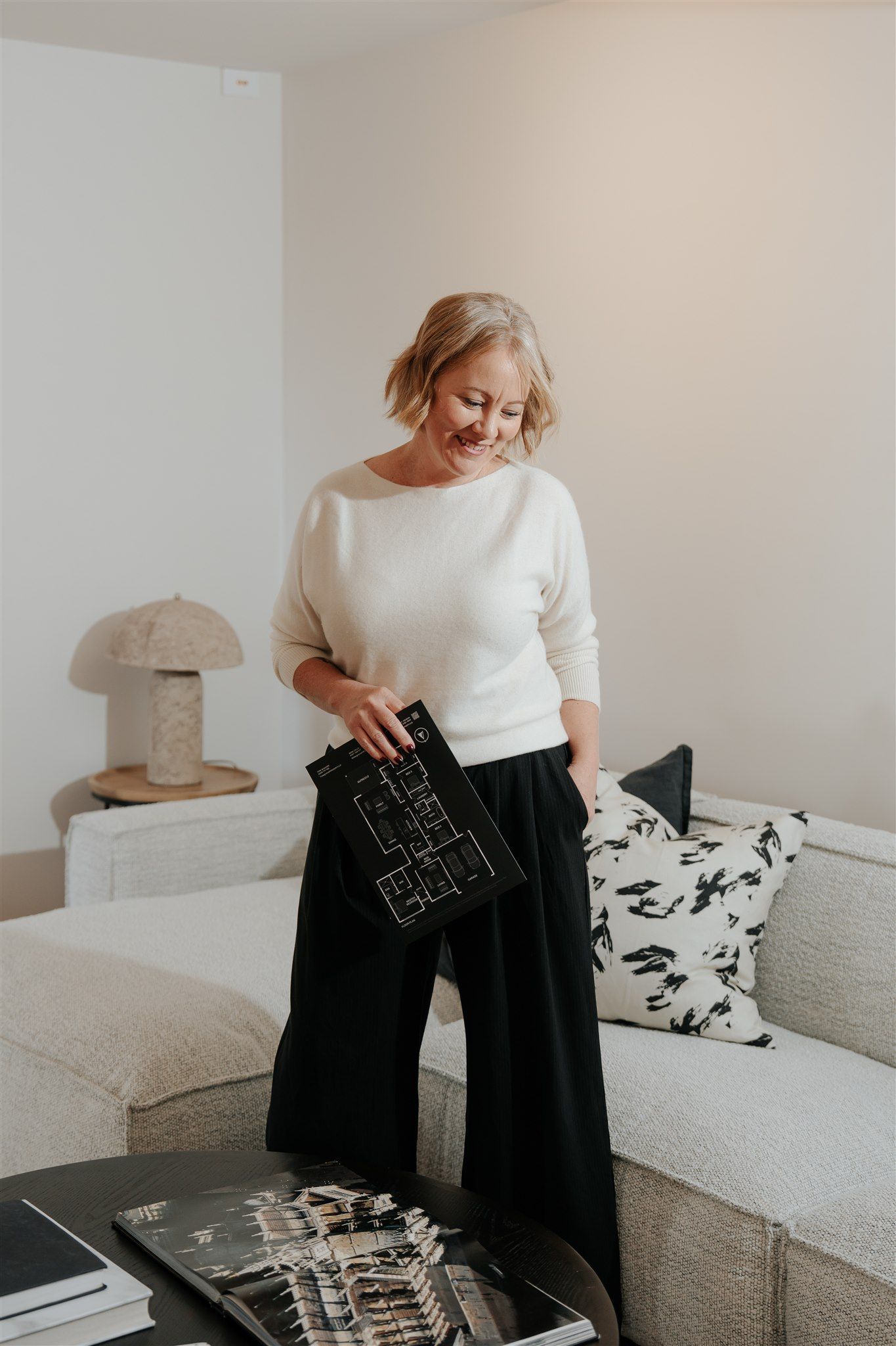 A woman is standing in a living room looking down and smiling — Hyde Burchell Buyers Agents In Canberra, ACT