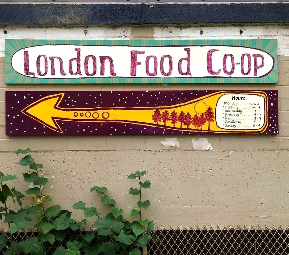 2 painted signs for the London Food Co- op with an arrow and purple trees with open hours listed.
