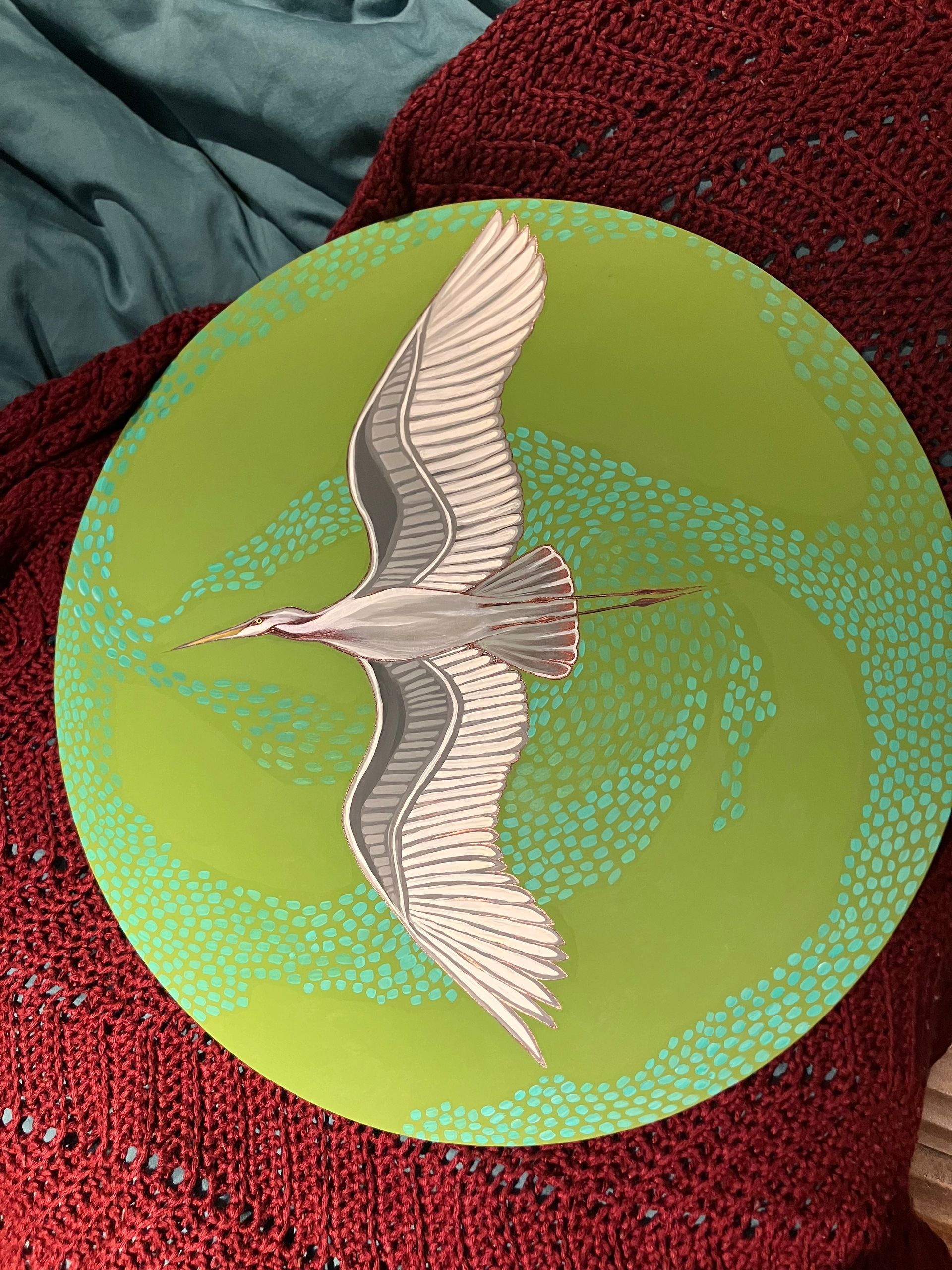 Image of a white and grey Egret flying, wings outstretched, riding the thermals with various shades of green in the background.