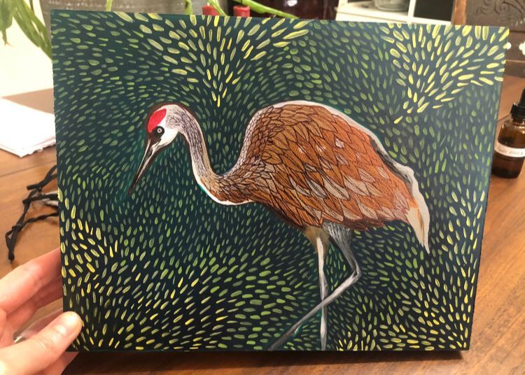 Painting of a sand hill crane with green plant leaves in the background.