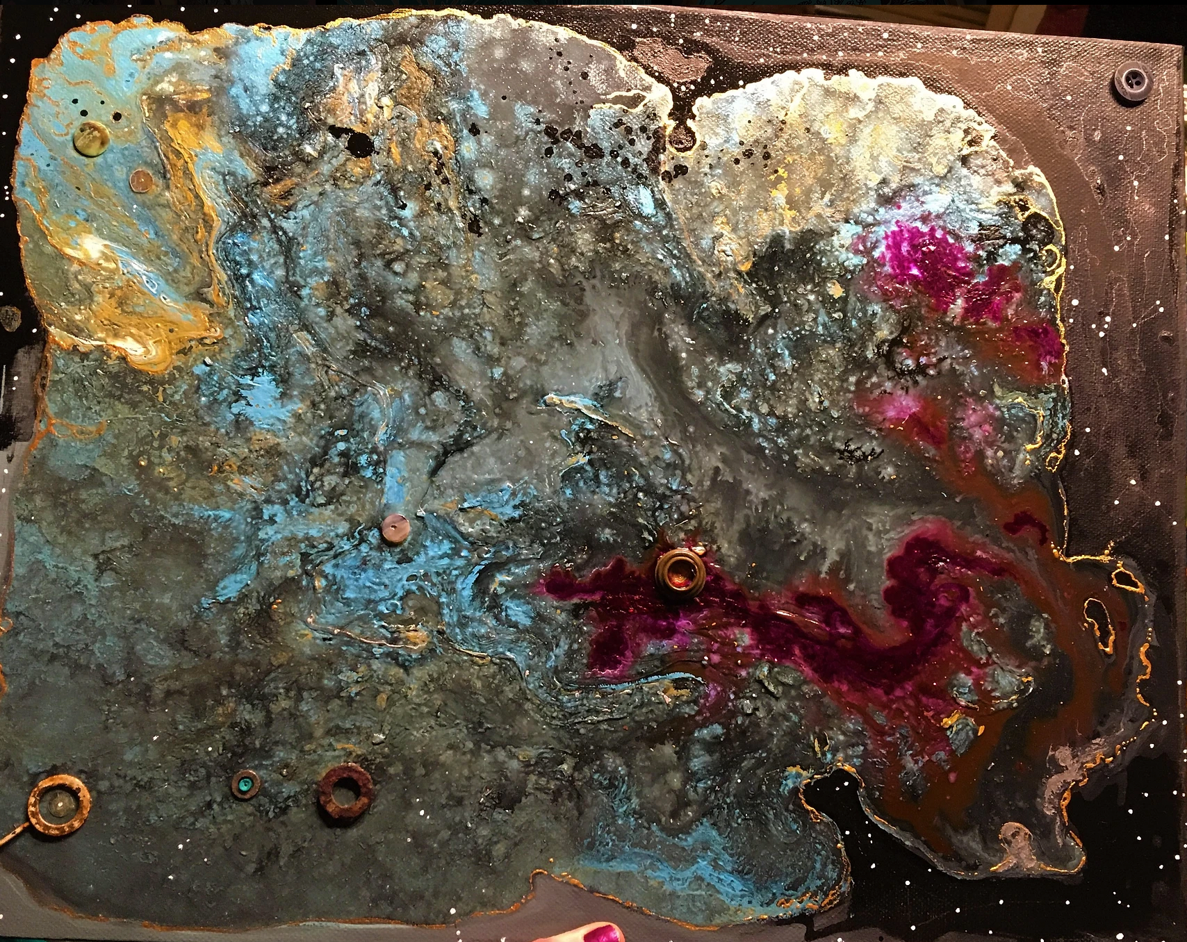 Image of a messy paint pour that looks akin to a galaxy. Hues of blue, burgundy, gold, black, adorned by some little metal pieces.