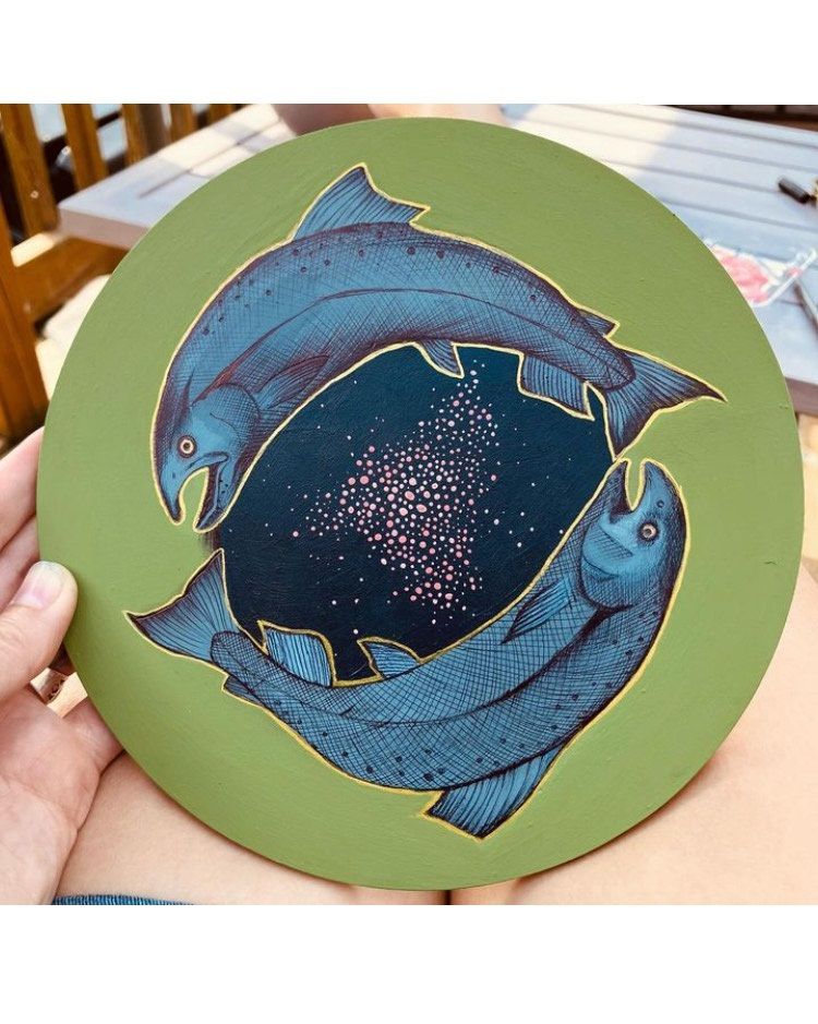 Image of two salmon swimming in the Pisces configuration, one with her head at the others tail and vice versa. Both Salmon are blue/grey with a galaxy of pink eggs between them , painted on a round wooden canvass with a light green background.