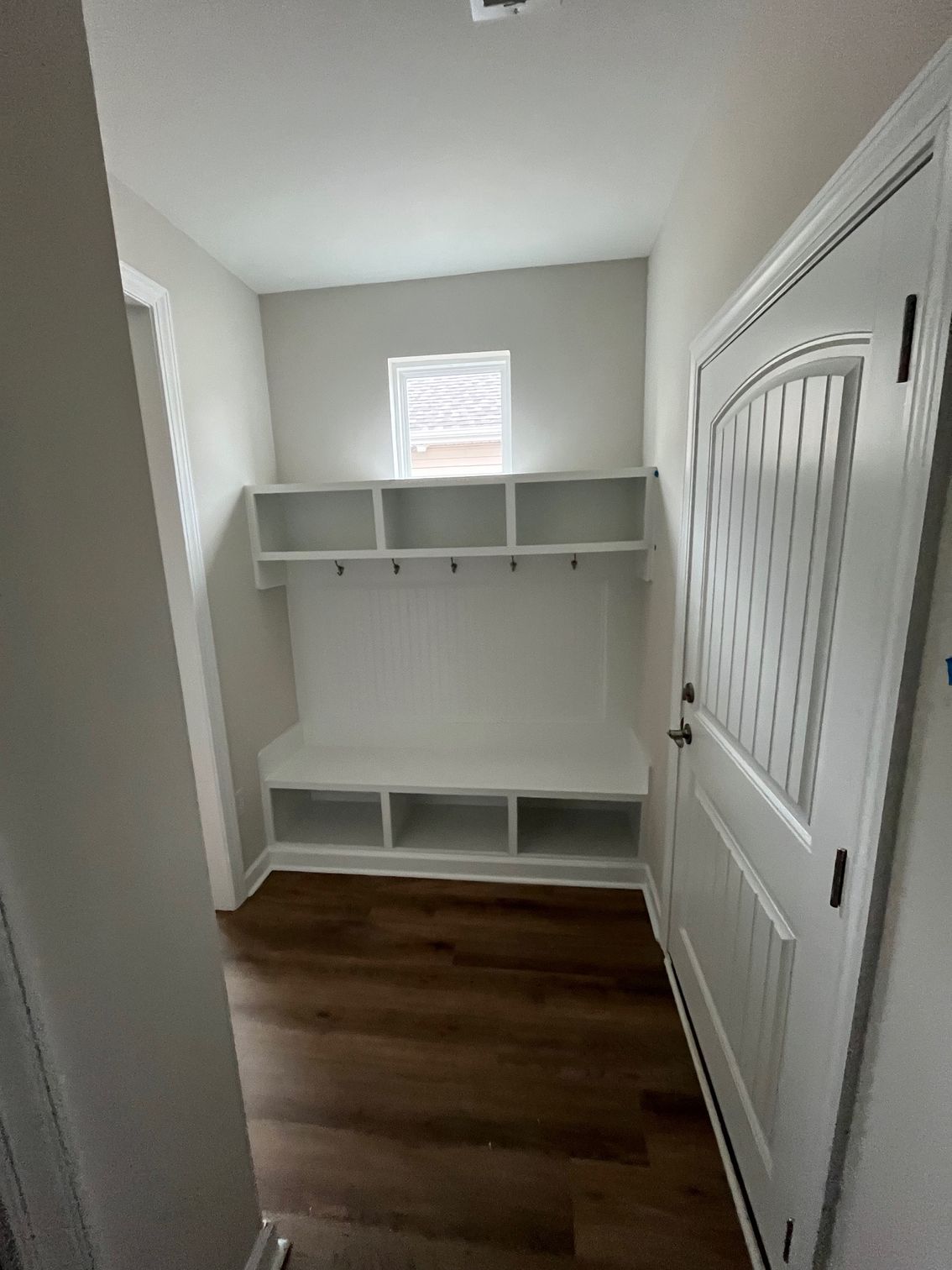 White mudroom with built-in bench, cubbies, and coat hooks, brown flooring, and a door.