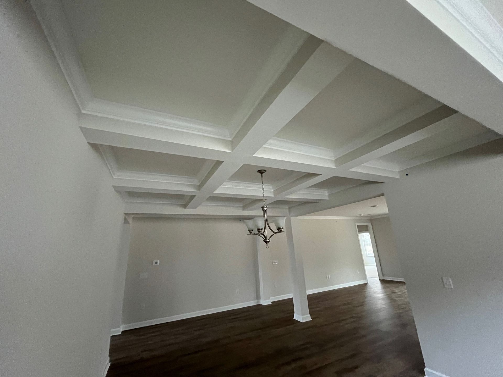 White coffered ceiling in a room with a chandelier, light walls, and dark wood floors.