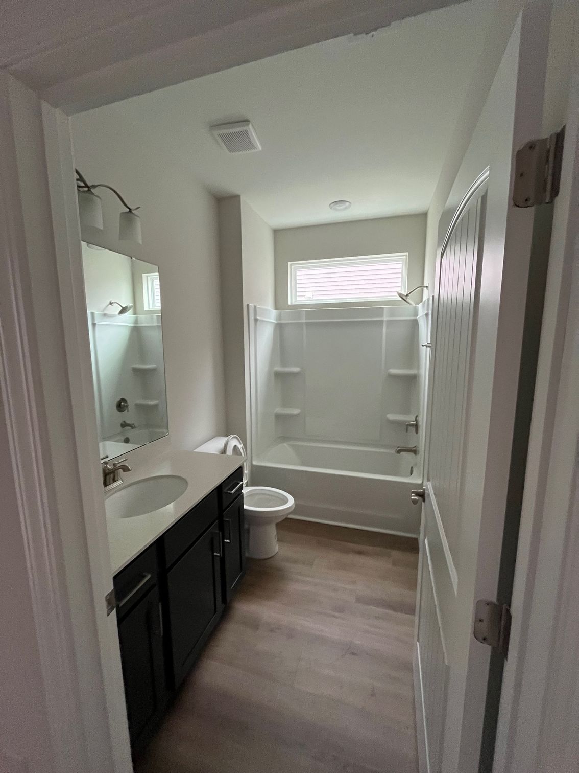 Bathroom with a dark vanity, white toilet, bathtub, and wood-look flooring.