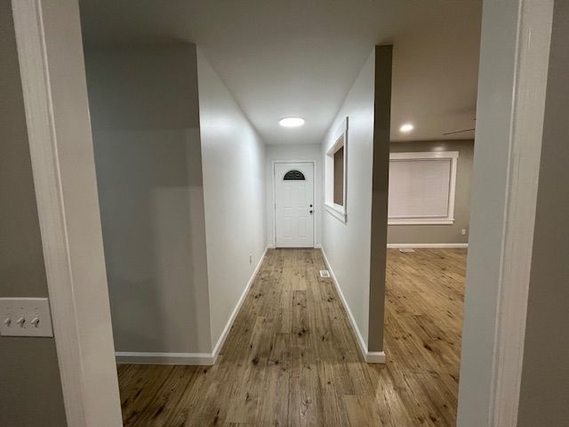 A hallway with wood-look flooring leads to a white door. A room with a window is visible on the right.