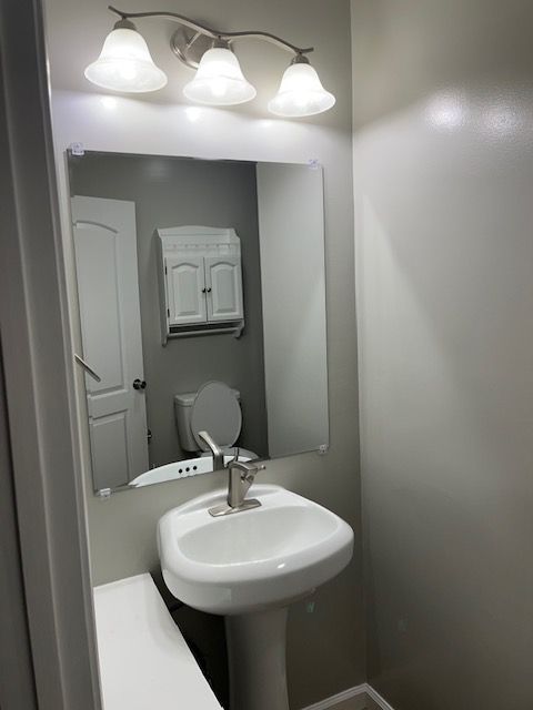 Bathroom with pedestal sink, mirror, and overhead lights. A cabinet is above the toilet.