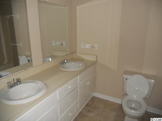 Bathroom with a white vanity with two sinks, a toilet, and a mirror. Beige walls and floor.