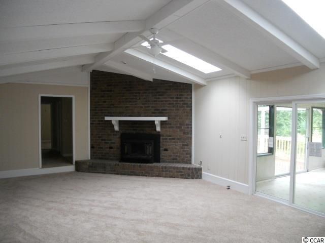 Empty living room with brick fireplace, beige walls, and carpet. Sliding glass door to sunroom.