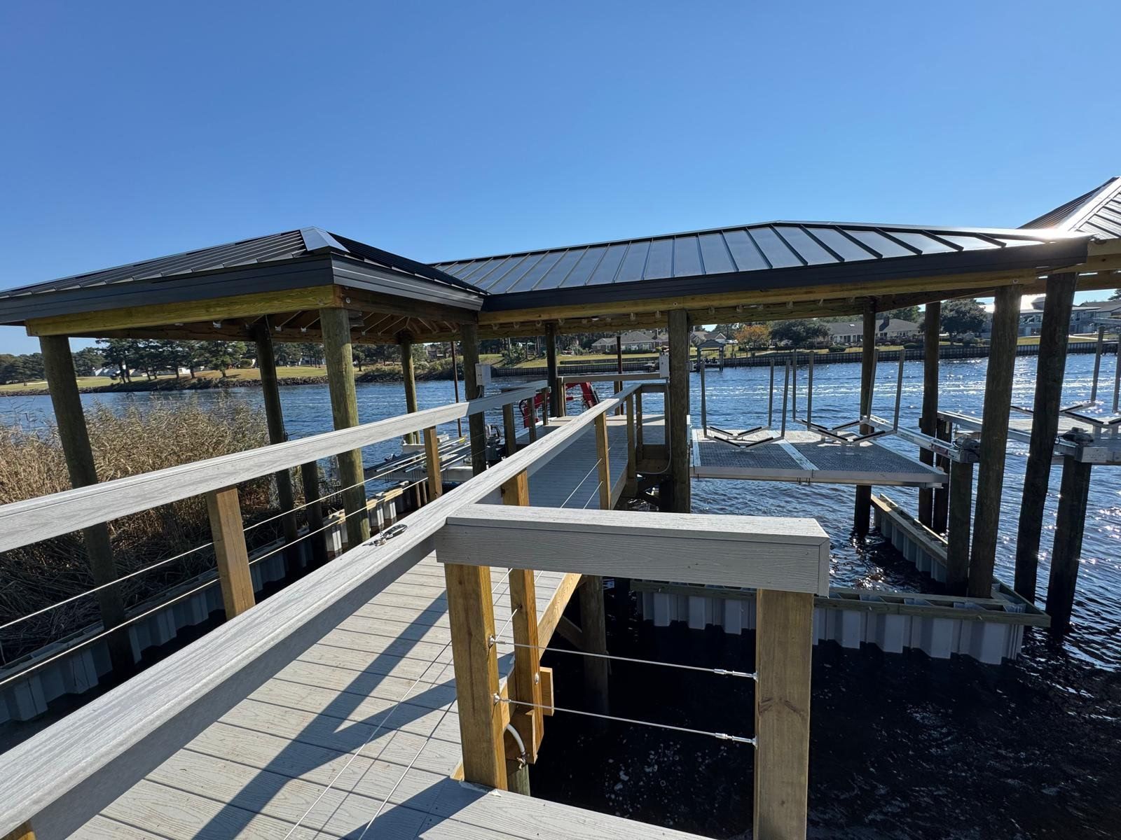 Wooden pier with shelter over water. Metal roof, cable railings, clear blue sky.