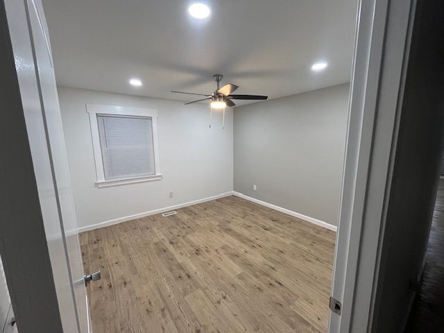 Empty bedroom with wood-look floor, window, and ceiling fan. Gray walls, recessed lights, and open doorway.