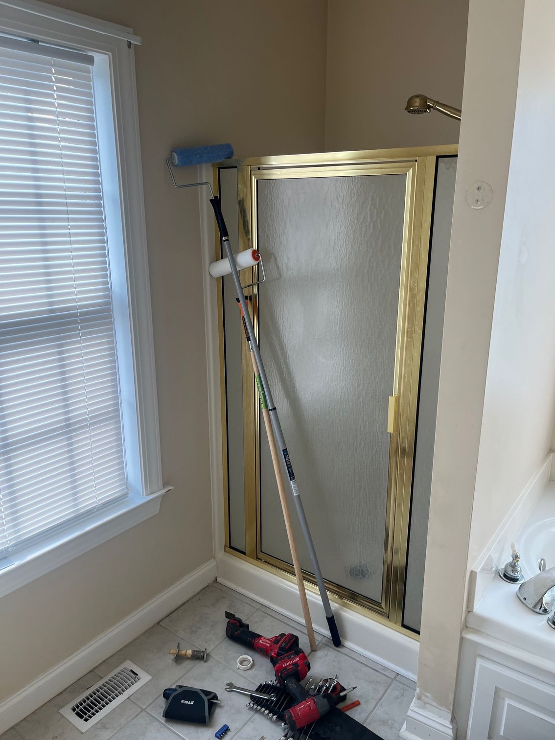 Bathroom with shower, window, and painting supplies. Roller and poles lean against the shower.