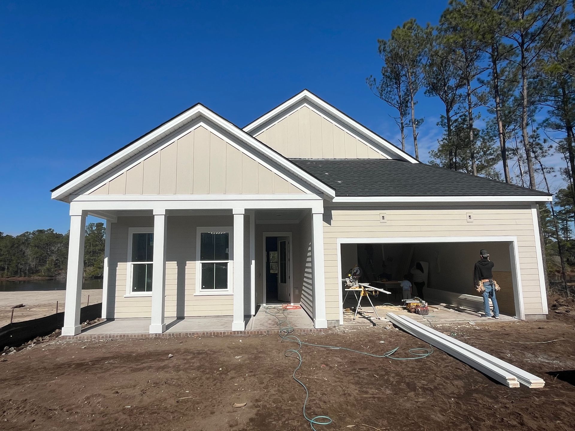 New house under construction with garage open, builders working outside. Tan siding, blue sky.