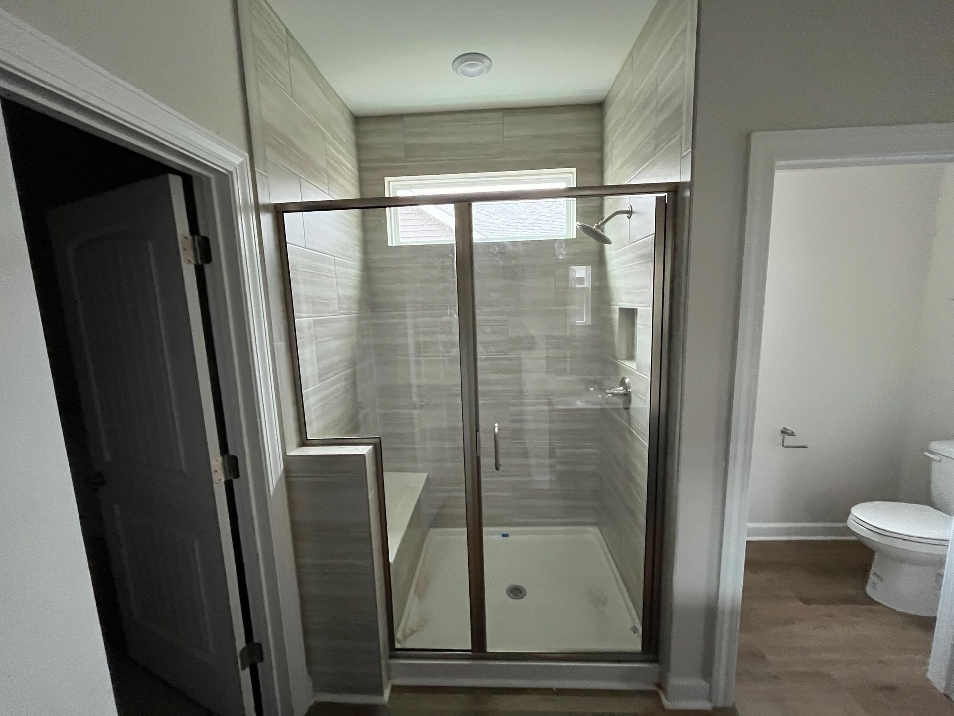 A shower with glass doors, built-in seat, and light gray tiled walls. A doorway is on each side.