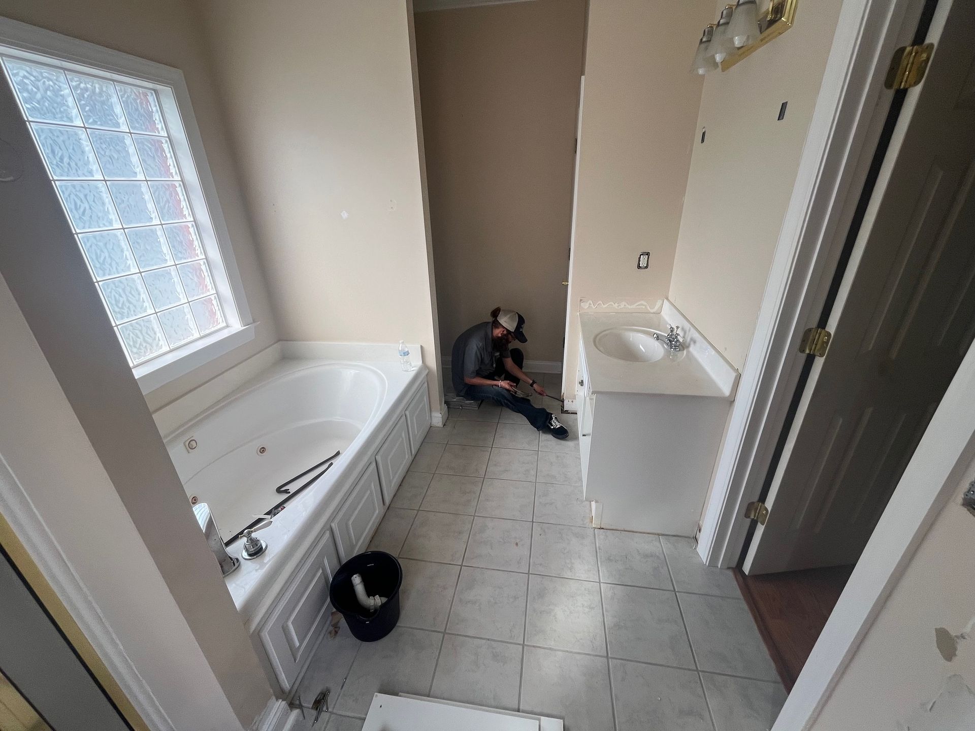 Bathroom renovation: worker on floor, tub, sink, window with grid, beige walls.