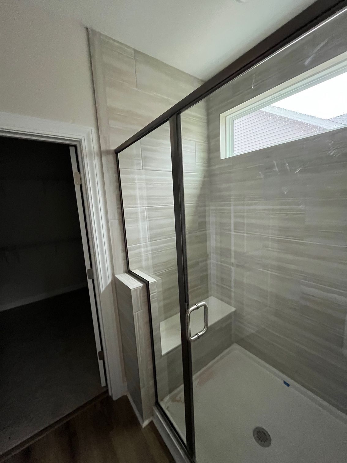 Shower with gray tile walls, dark frame glass door, built-in bench, and small window.