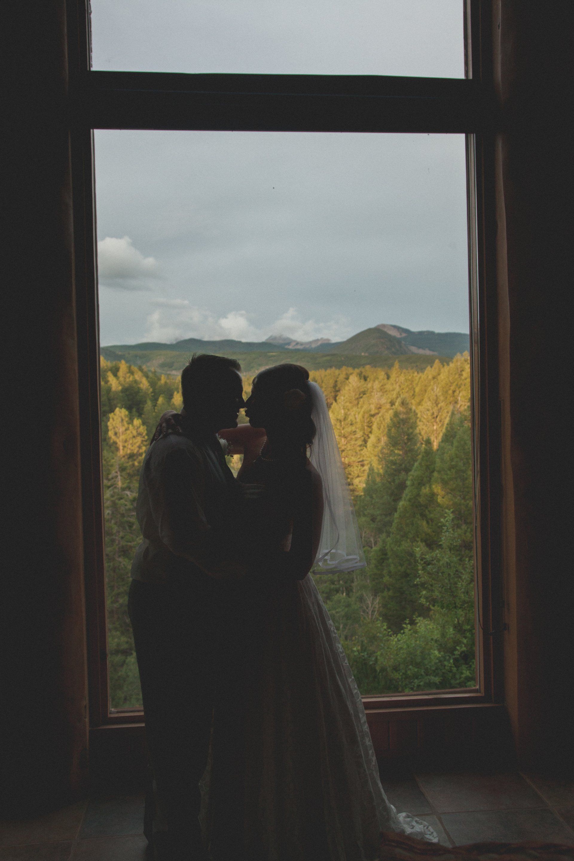 A bride and groom are kissing in front of a large window.
