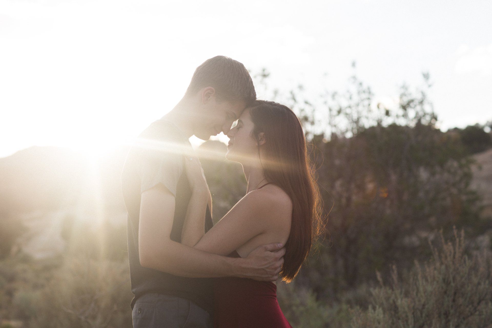 A man and a woman are kissing in a field with the sun shining through the trees.