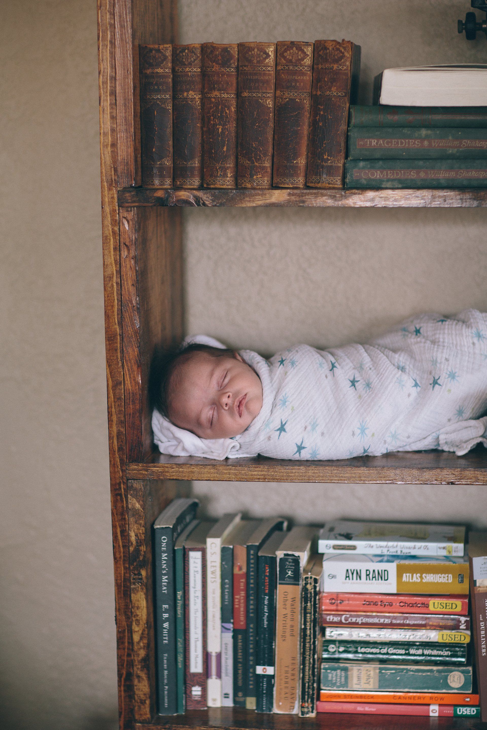 A baby wrapped in a blanket is sleeping on a bookshelf.