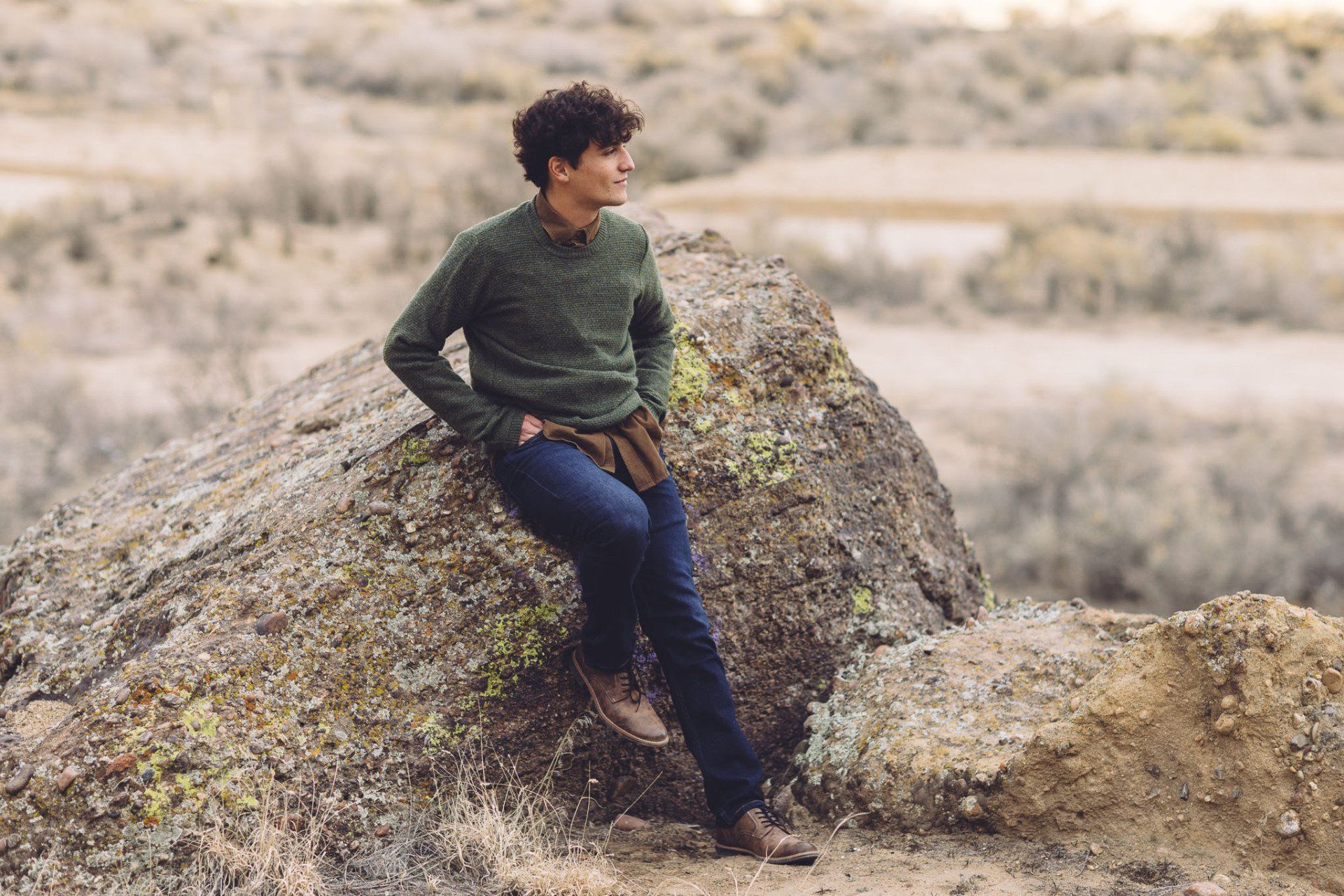 A young man is sitting on a large rock in the desert posing for senior pictures