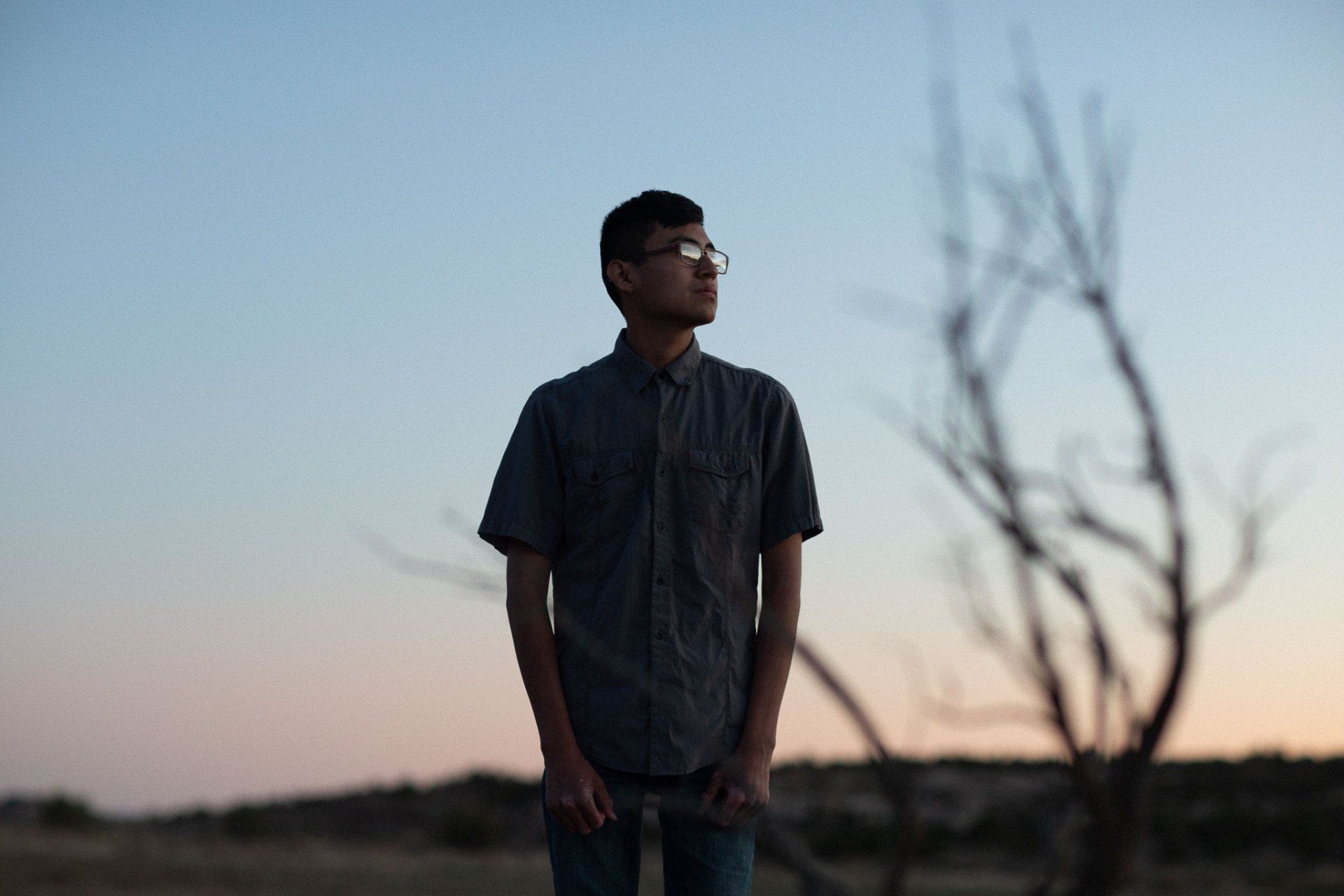 A young man wearing glasses is standing in front of a tree posing for senior pictures