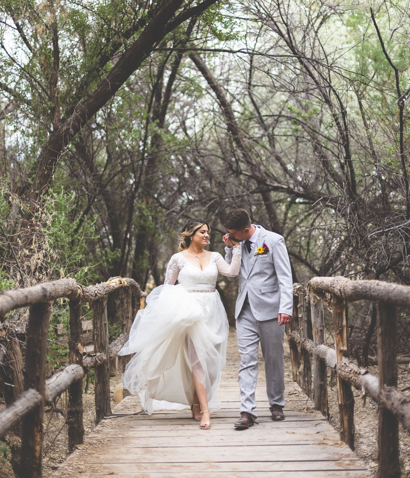 A bride and groom are walking across a wooden bridge in the woods.