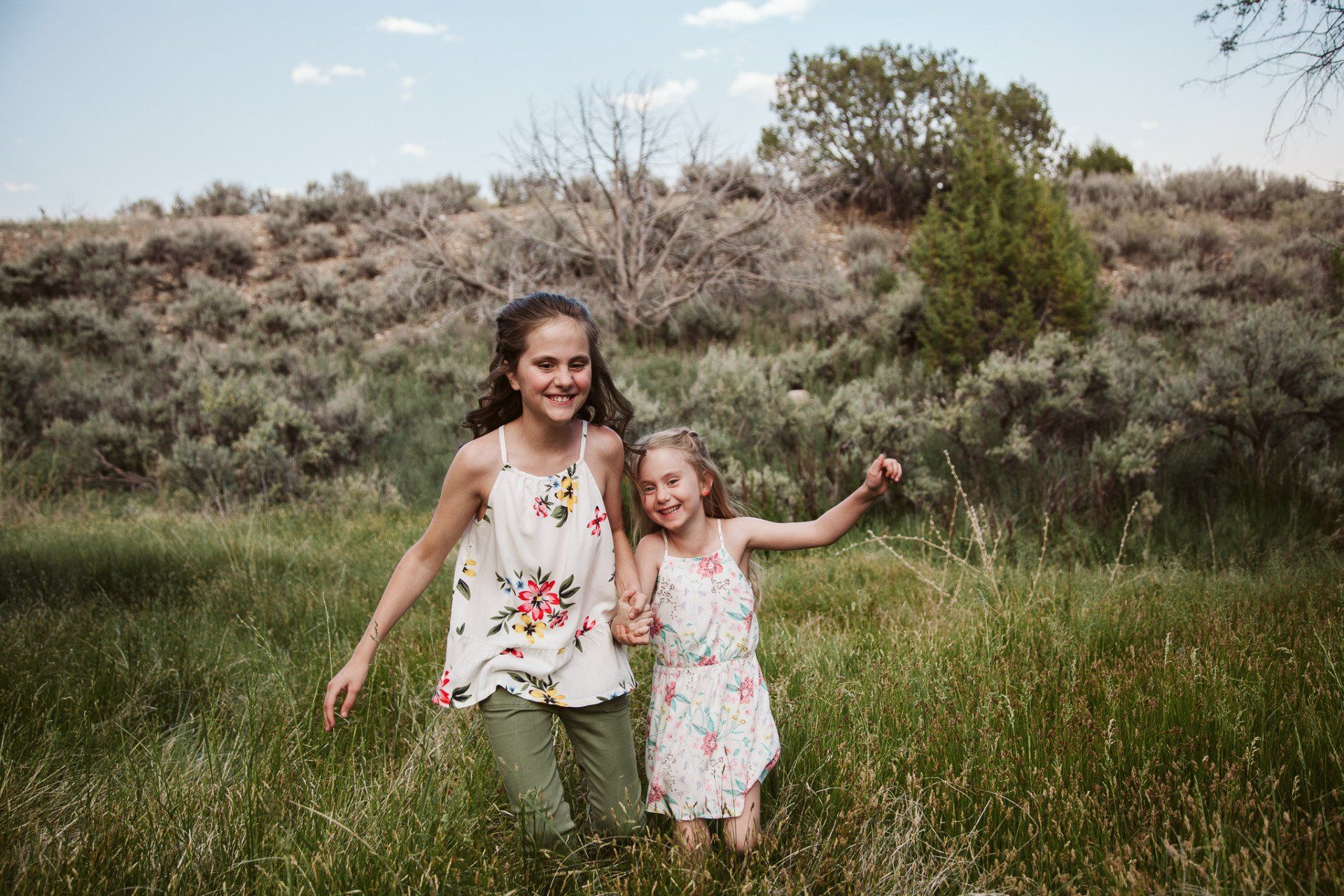 Two young girls are running through a grassy field holding hands.