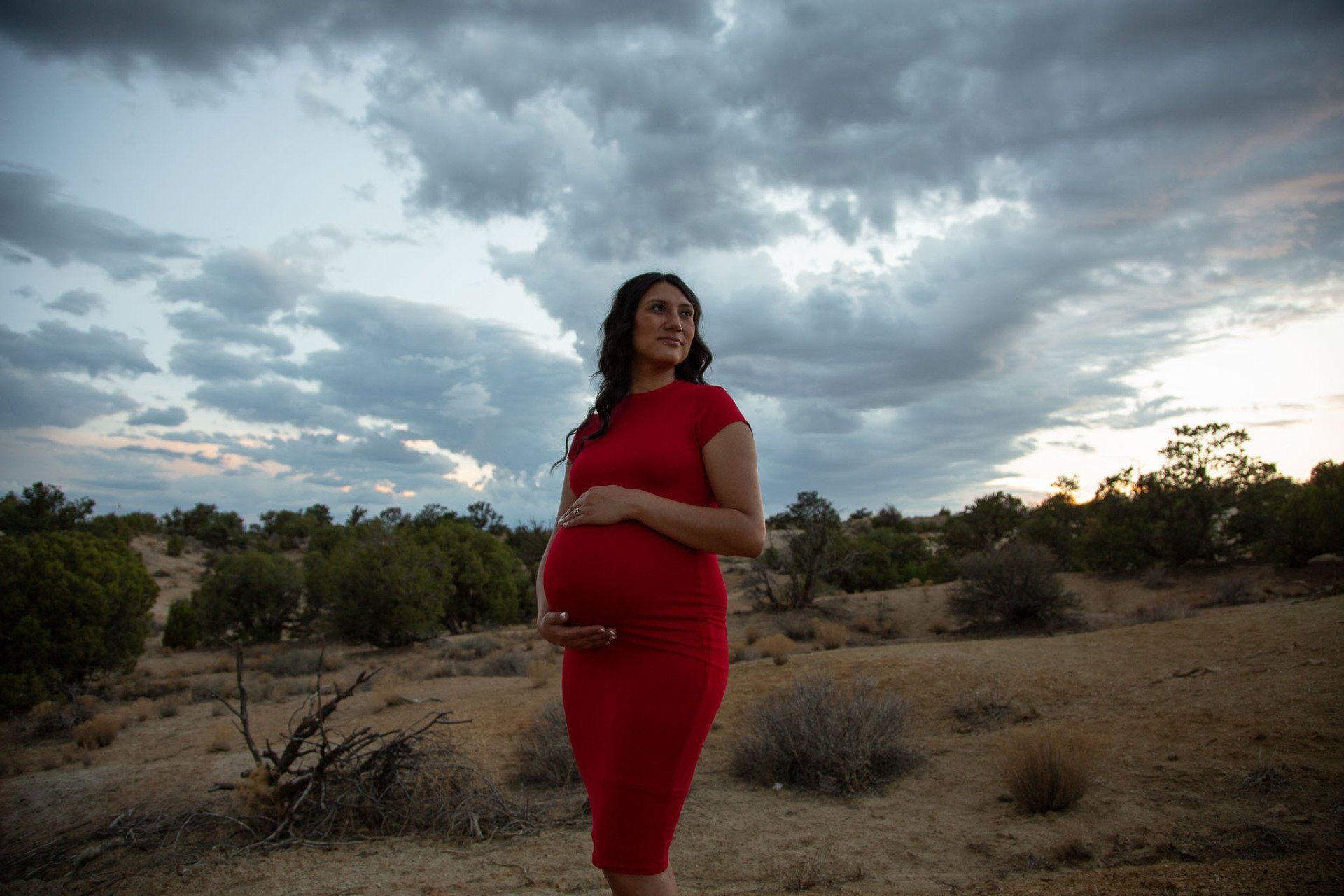 A pregnant woman in a red dress is standing in the desert holding her belly.